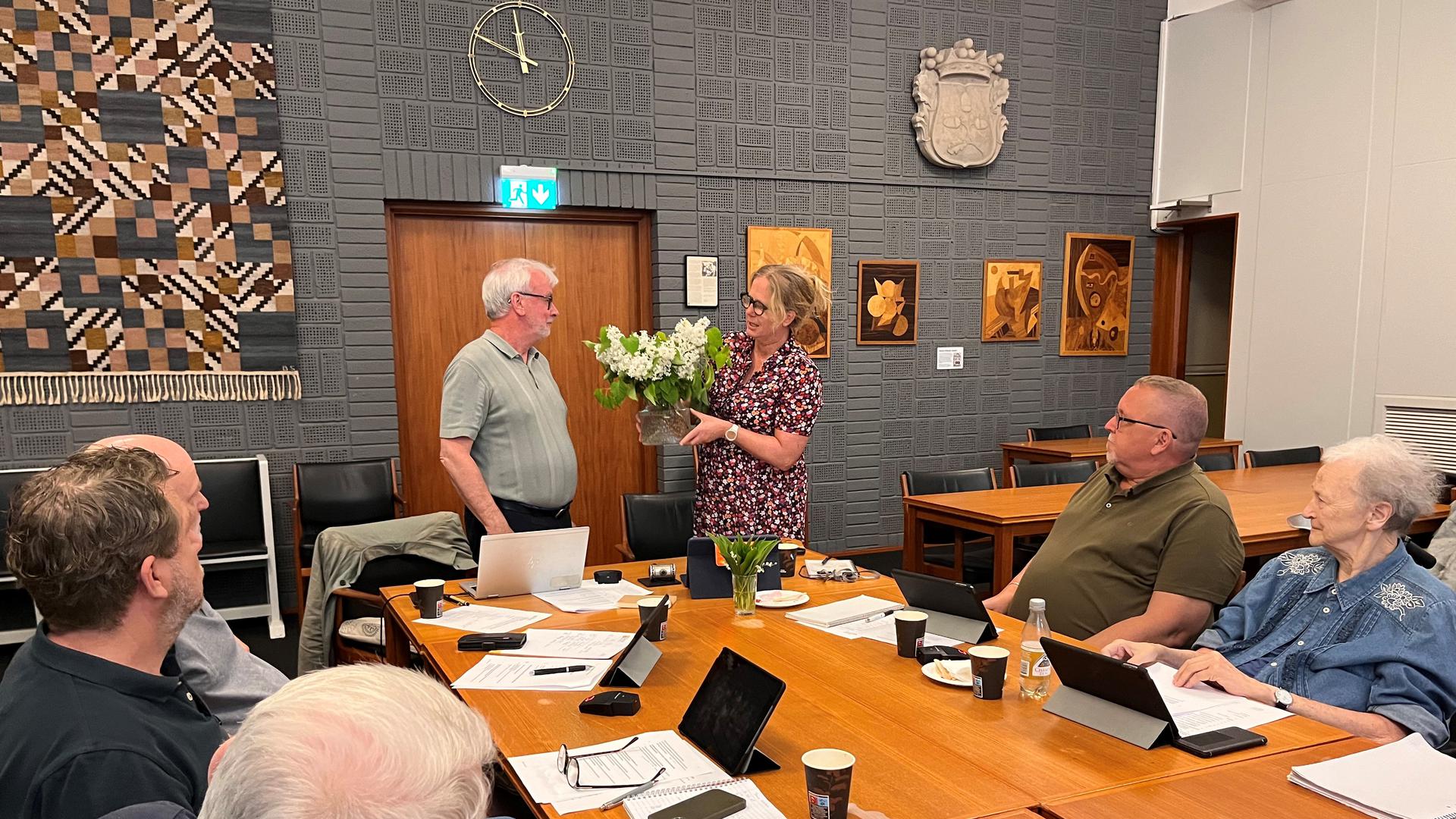 Sammanträdesrum med ledamöter som avtackar med en vit blomma i en vas